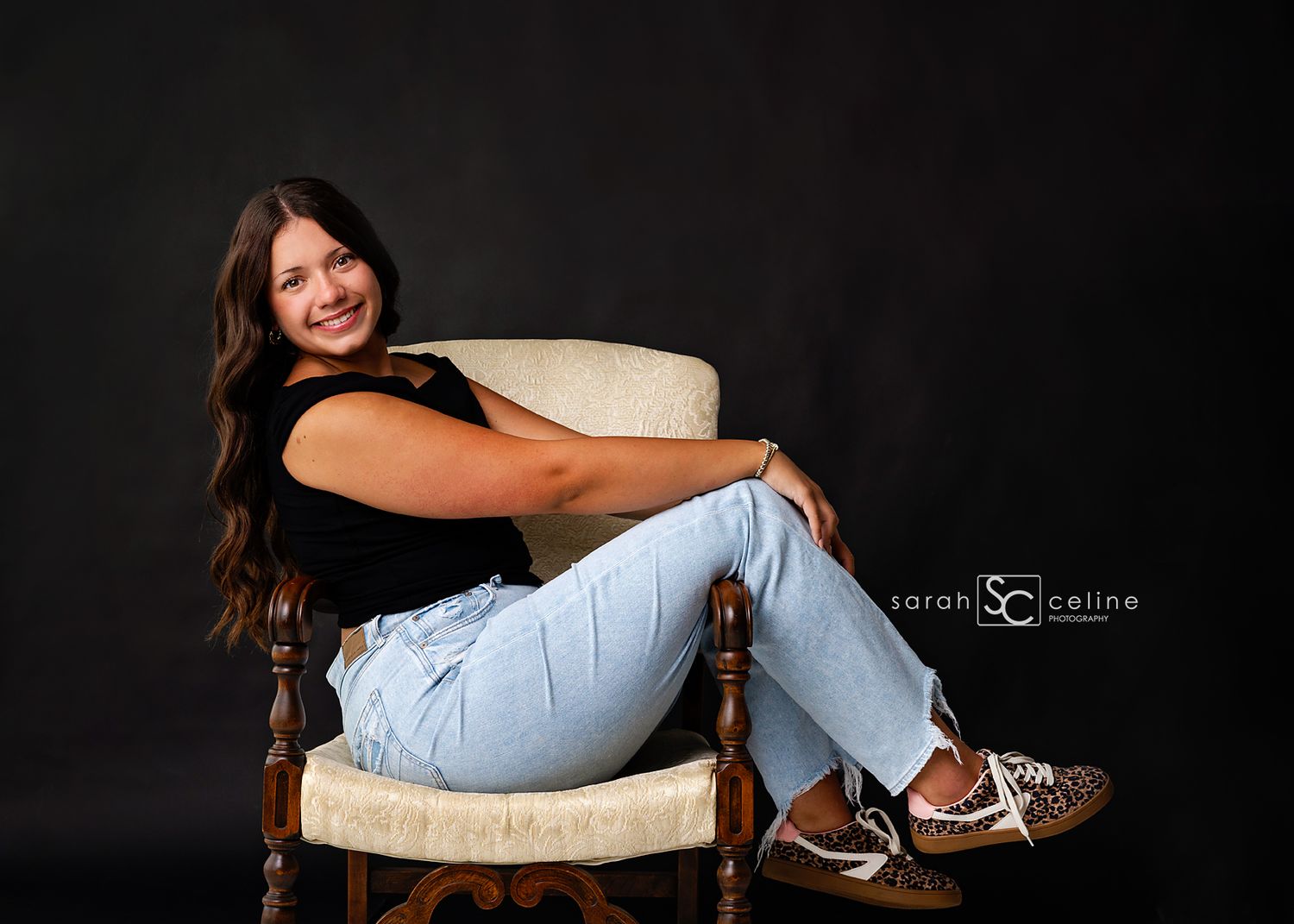 Young woman smiling in black top and light jeans, seated on vintage chair against dark background, professional portrait photo.