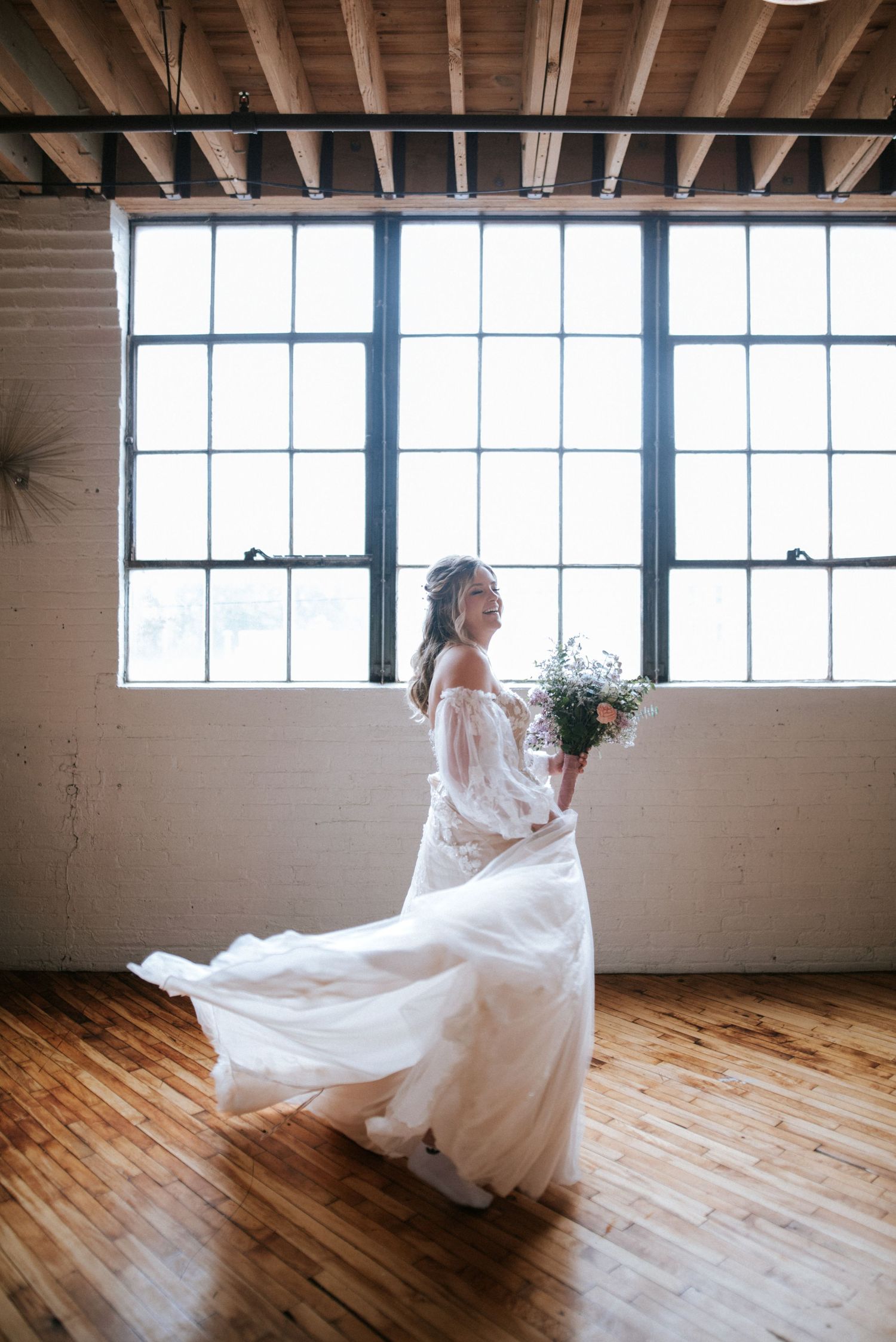 Bride in flowing white off-shoulder gown twirling with bouquet in sunlit industrial loft with hardwood floors.