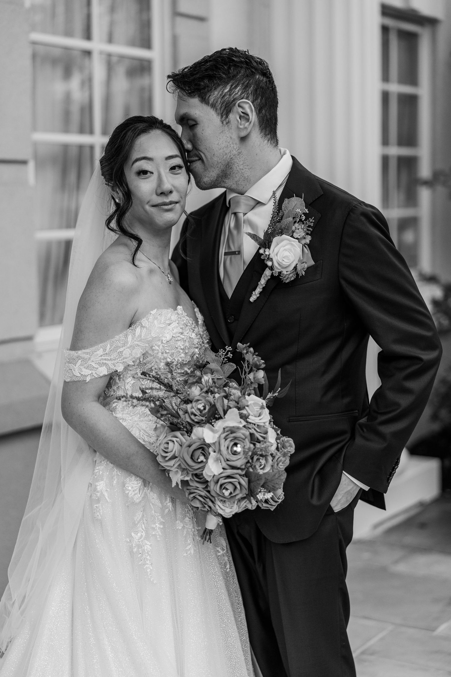 Black and white wedding photo of a groom kissing bride on the cheek, bride holding a rose bouquet.