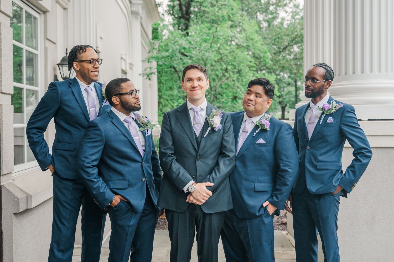 Groom in gray suit stands with four groomsmen in navy blue suits and floral boutonnieres outside a white columned building.