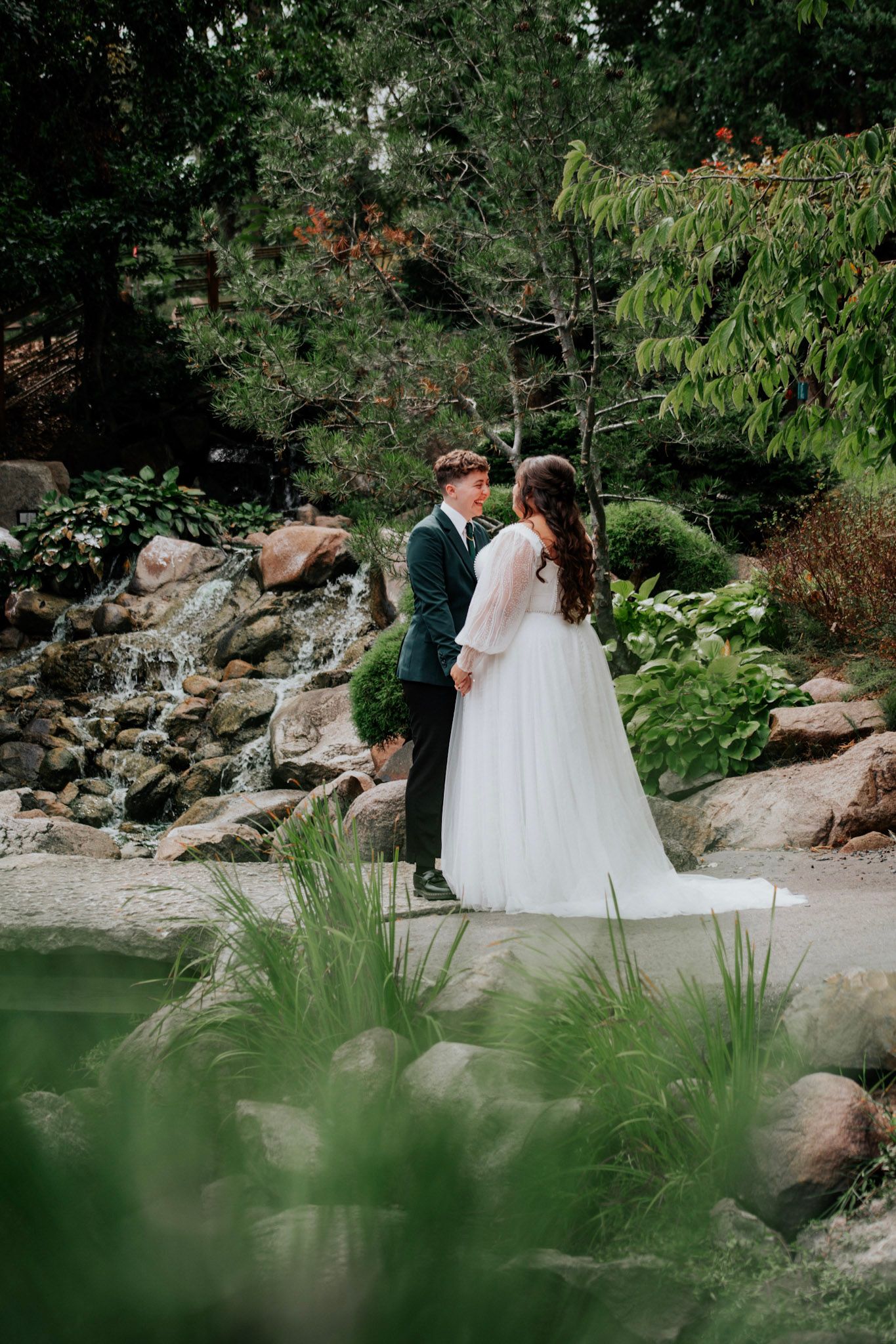 Bride and groom hold hands near a serene garden waterfall surrounded by lush green landscaping.
