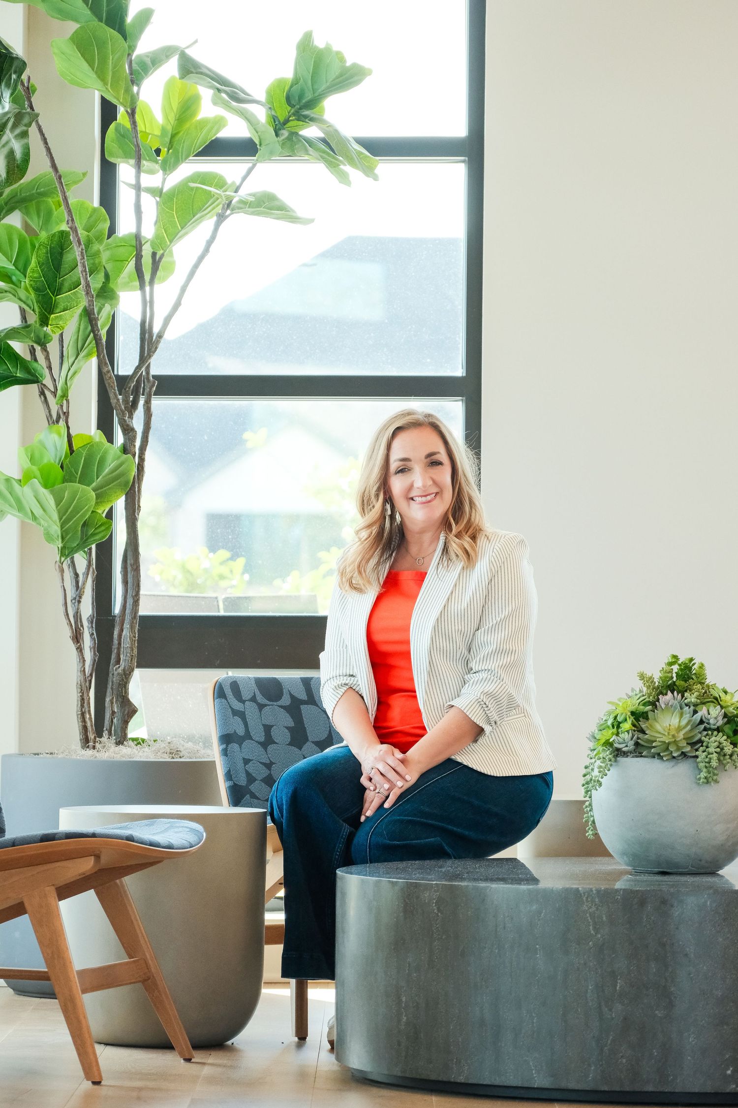 Smiling woman in striped blazer and red top leaning on wooden counter in modern interior space.