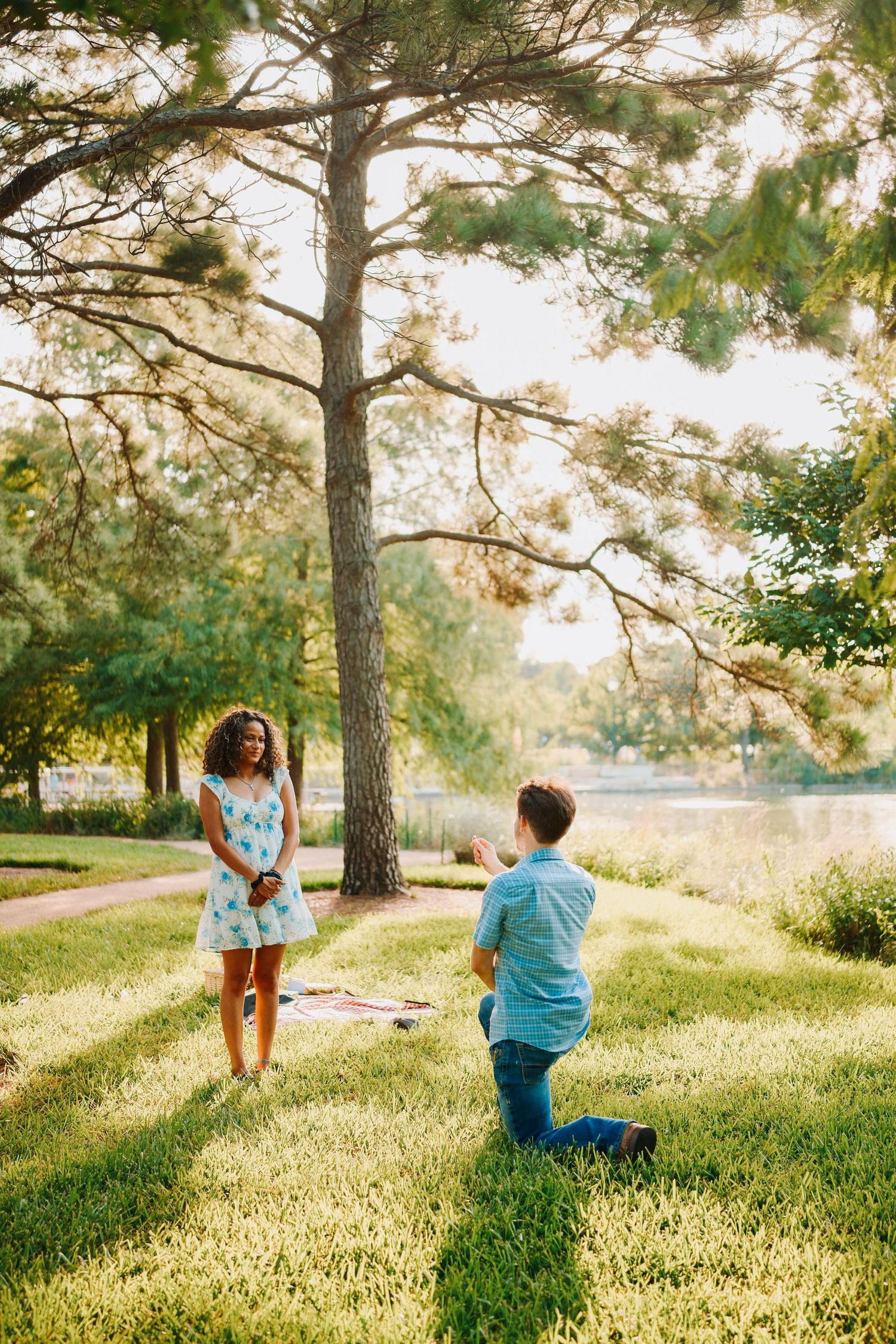 Man kneeling on one knee proposing to woman in blue dress in a sunlit park near a lake.