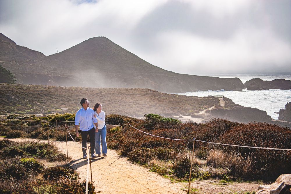 Couple walking on coastal trail with rocky shoreline, misty hills, and dramatic ocean views in background.