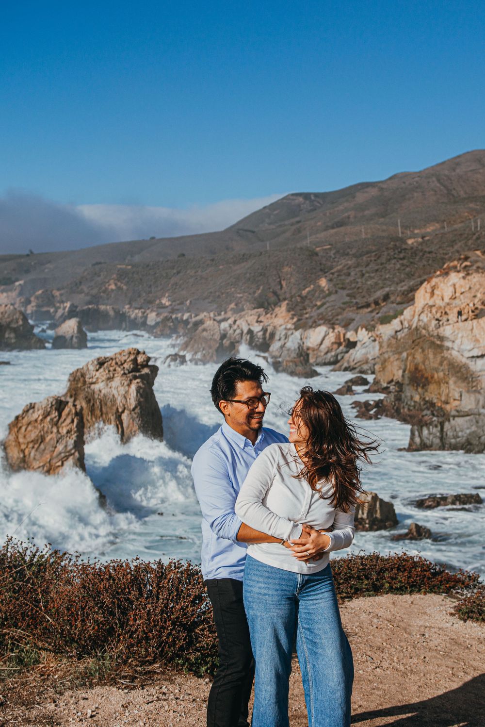 Couple embracing on rocky Big Sur coastline with crashing waves and mountains under a clear blue sky.
