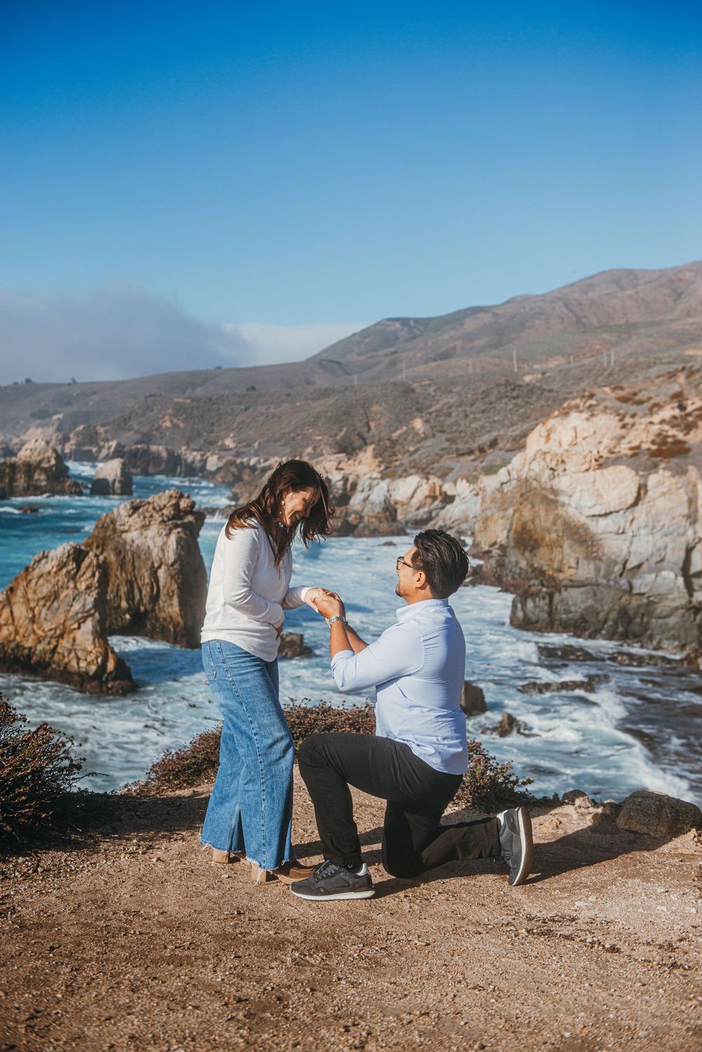 Man proposing to woman on rocky coastal cliffside with ocean waves and mountains in background on sunny day.