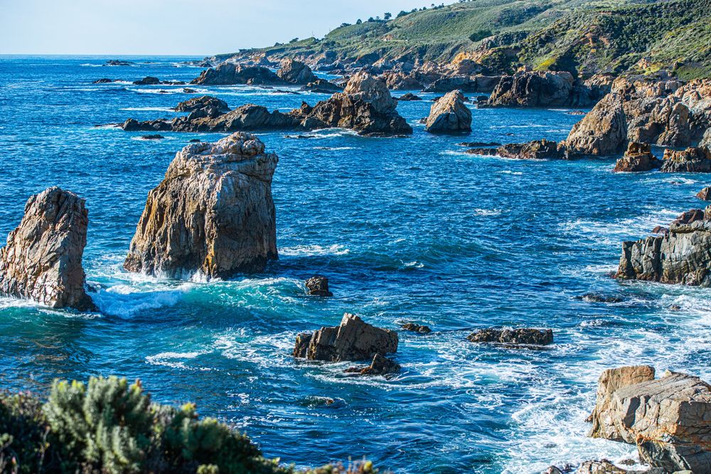 Rocky coastal seascape with jagged stone formations rising from vivid blue ocean waves along a rugged green clifftop shoreline.