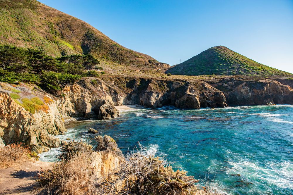 Scenic Big Sur coastline with turquoise waves, rocky cliffs, lush green hills, and a cone-shaped mountain under clear blue skies.