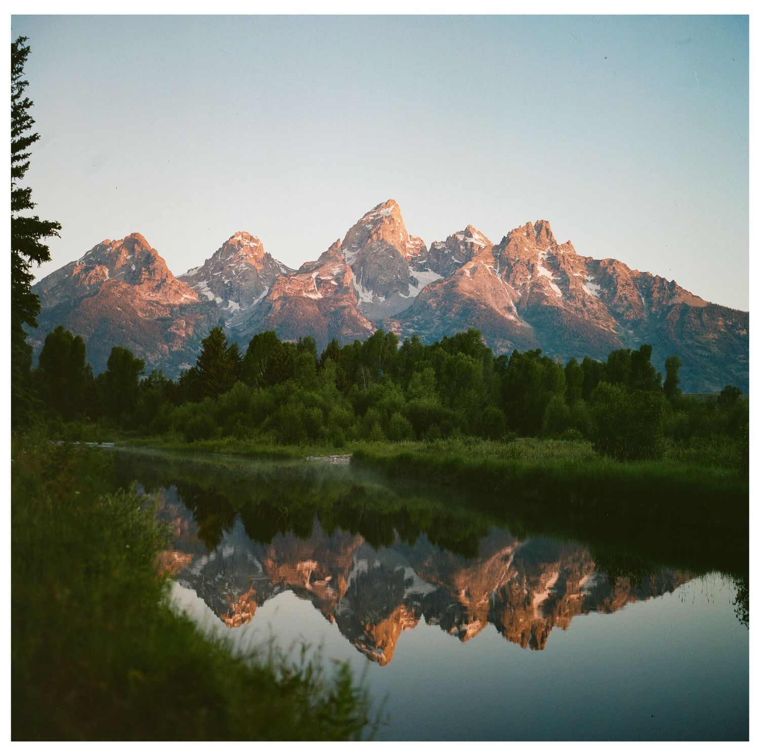 Grand Teton mountains reflect perfectly in still river waters at golden sunrise.