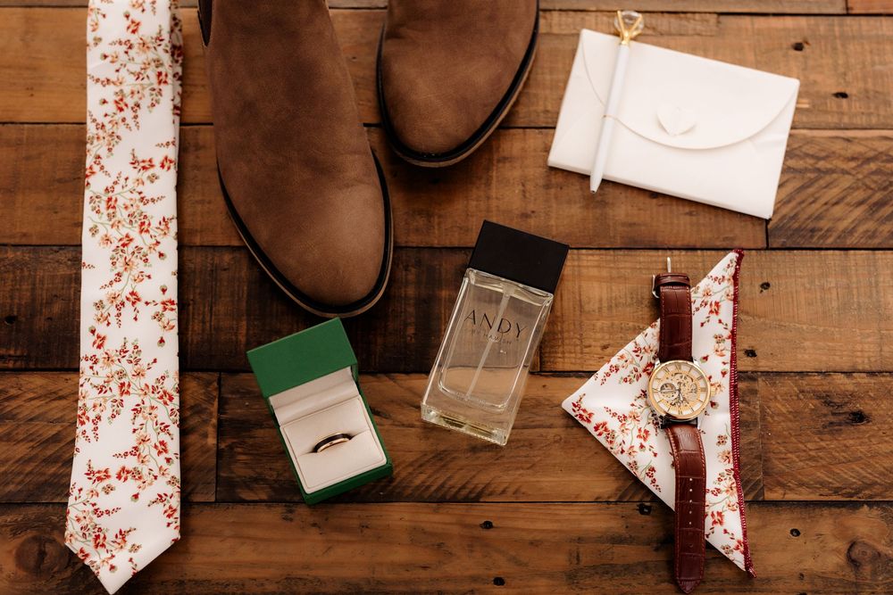 Groom wedding flat lay with floral tie, brown boots, ring box, cologne, watch, pocket square, and white wallet on wood.