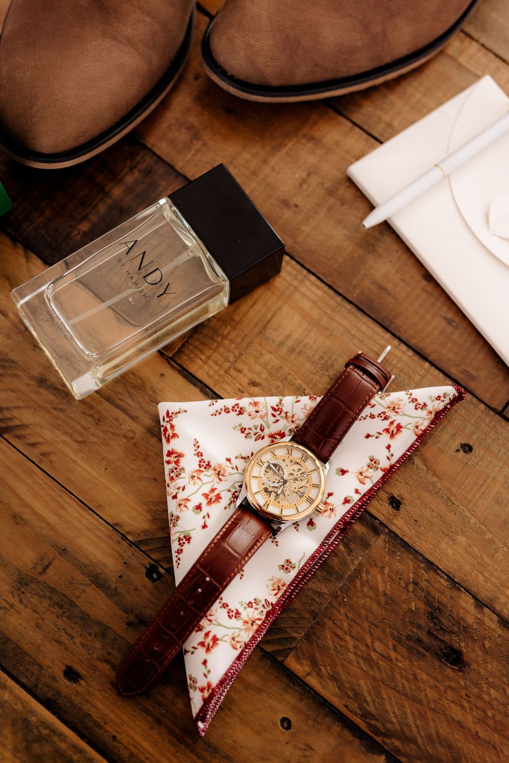 Groom accessories flat lay with brown leather watch, floral pocket square, cologne, shoes on rustic wood.