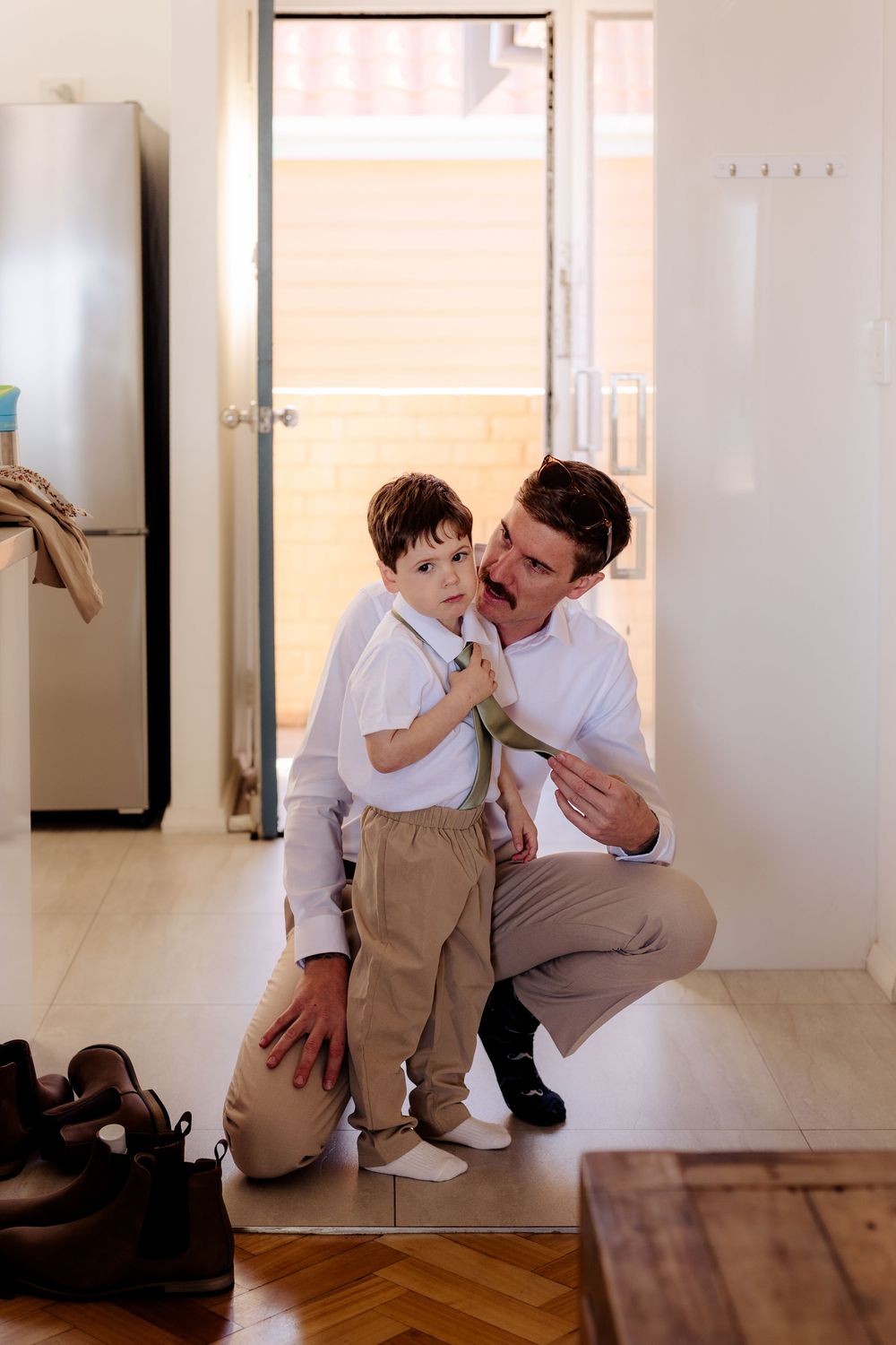 Father crouching down to help young son adjust tie, both dressed in white shirts and khaki pants at home.