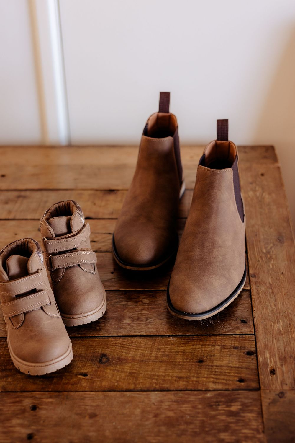 Two brown leather boots, one small child's shoe and one adult Chelsea boot, displayed on a rustic wooden surface.