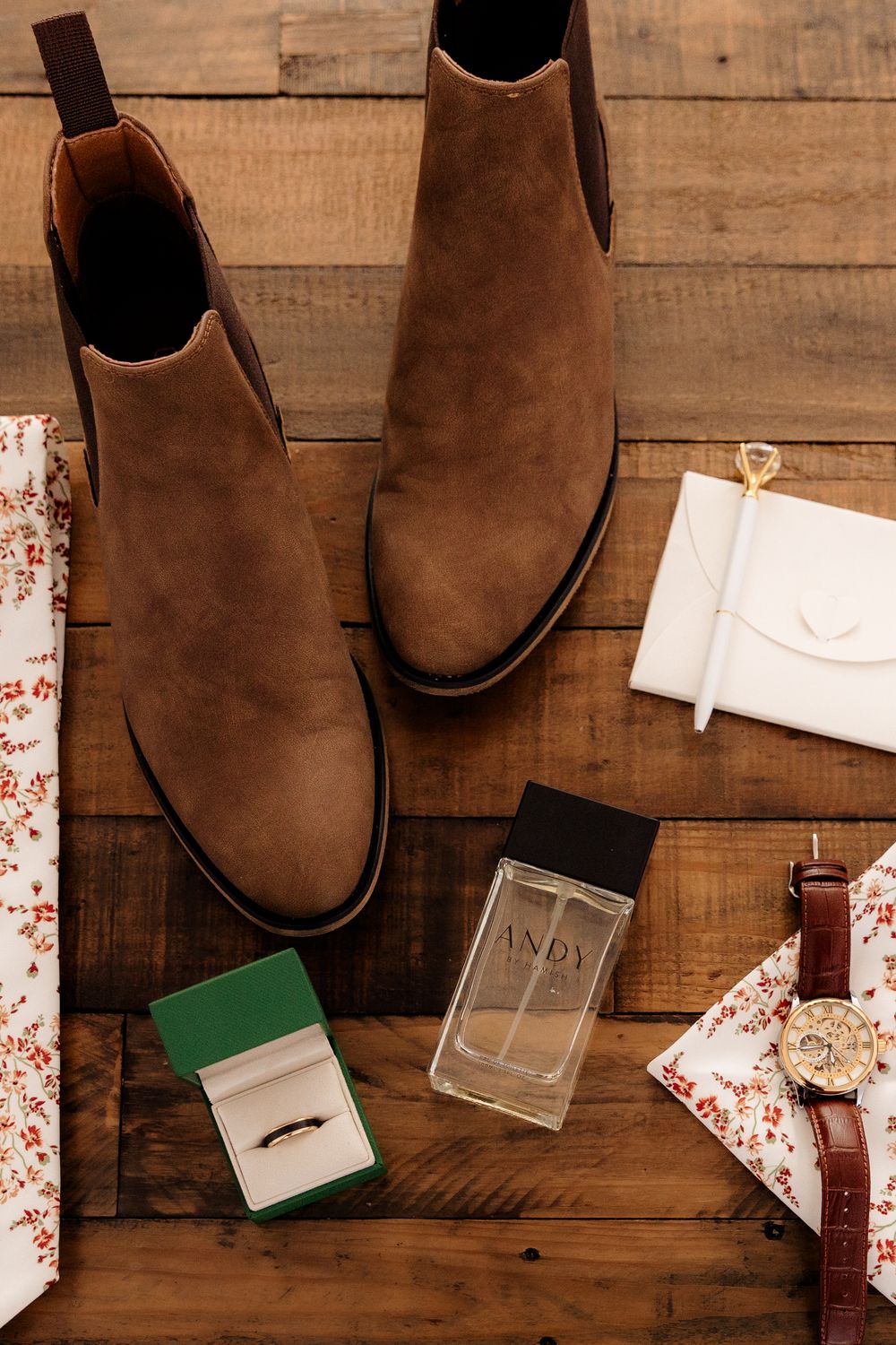 Brown suede Chelsea boots with cologne, wedding ring, watch, and floral accessories arranged on rustic wooden floor.