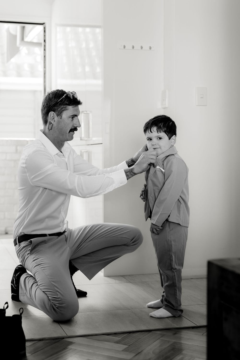 Black and white photo of a father kneeling to adjust his young son's suit jacket indoors.