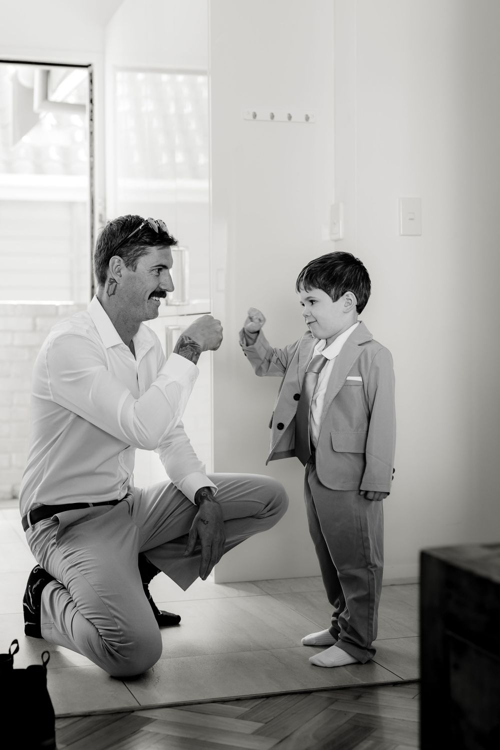 Man in dress shirt fist bumps young boy in suit, sharing a tender moment before a wedding ceremony.
