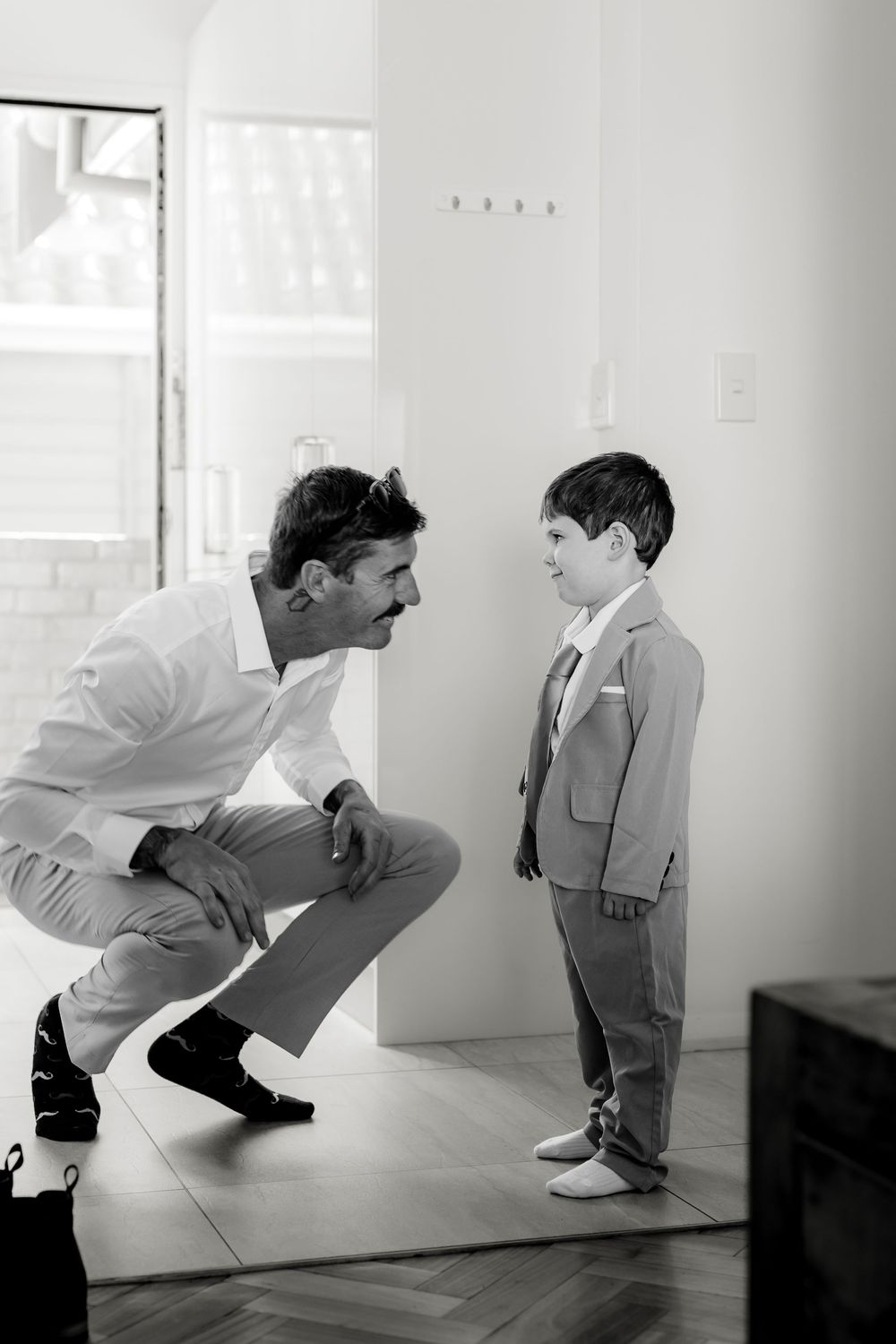 Black and white photo of a father crouching down to speak with his young son dressed in a suit.