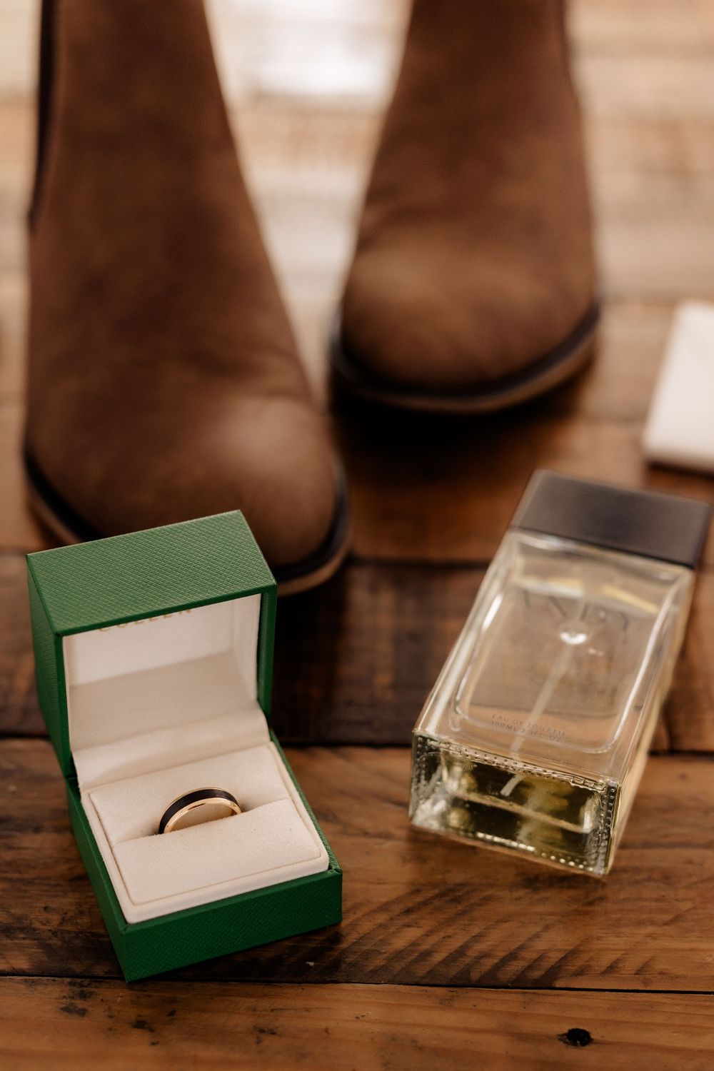 Gold wedding ring in green box beside cologne bottle and brown leather boots on wooden floor.