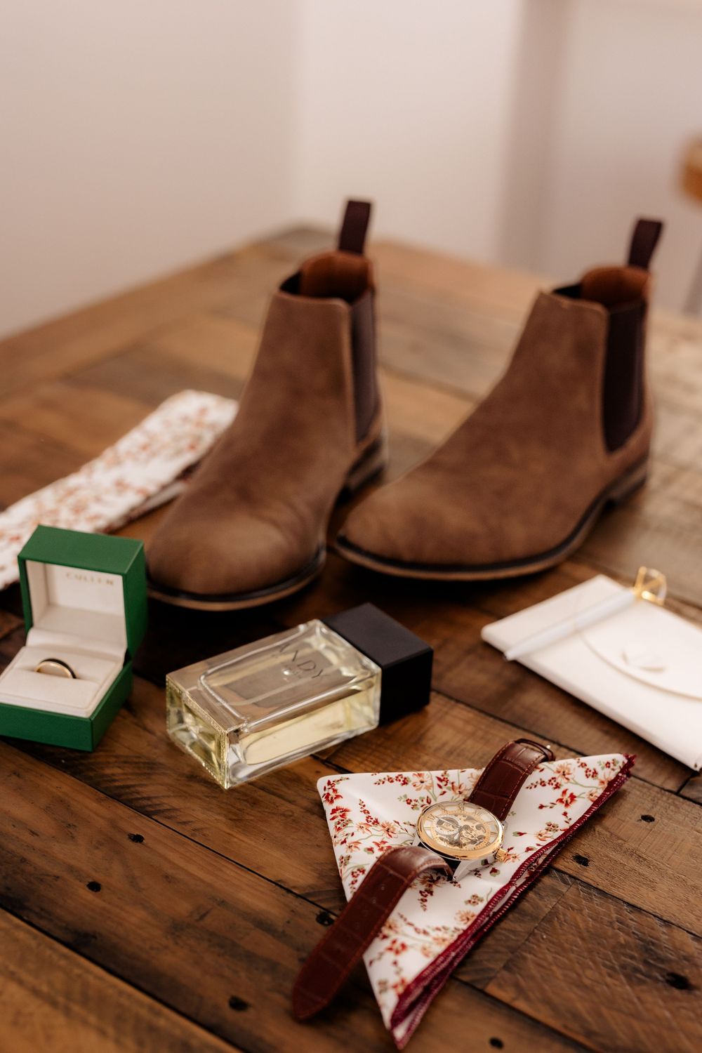 Brown Chelsea boots with cologne, a ring box, floral bow tie, watch, and pocket square arranged on a rustic wooden table.