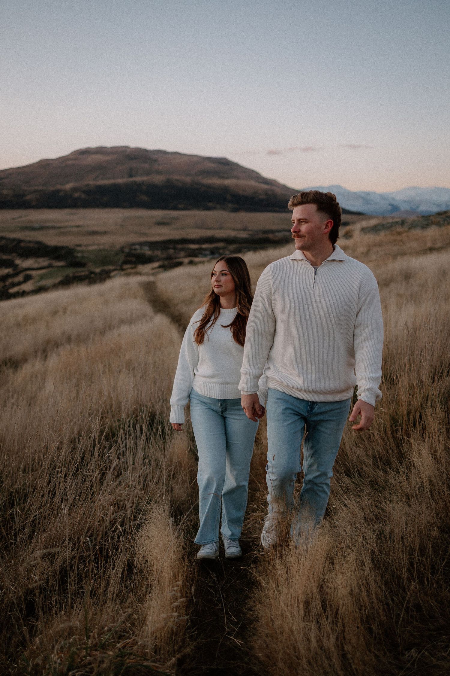 Couple in white sweaters and jeans walking through golden grass fields with mountain backdrop at dusk.