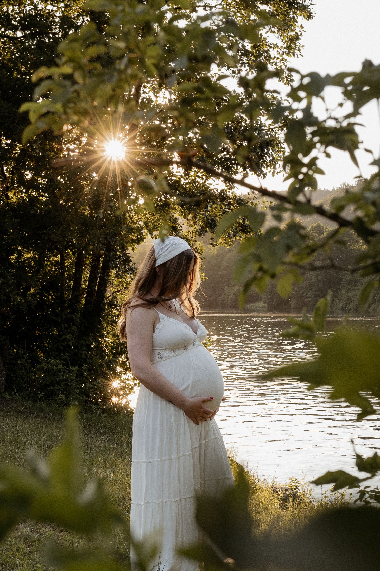 Pregnant woman in white dress holding belly by sunlit lake at golden hour, framed by lush green trees.