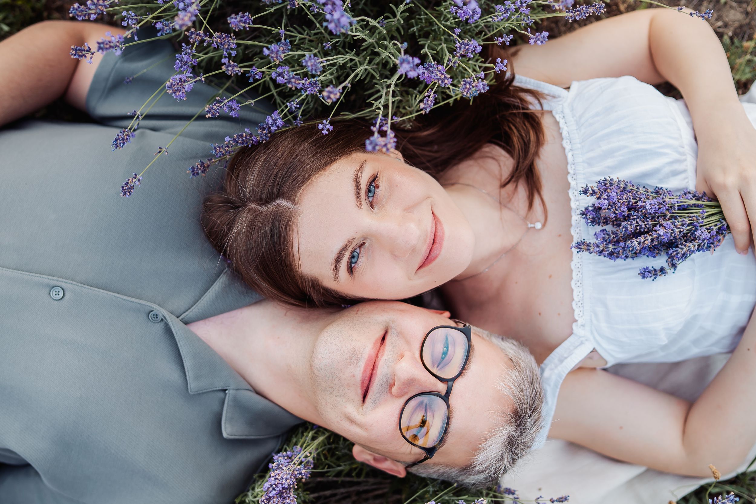 Couple lying in lavender field, young woman resting head on older man with glasses, surrounded by purple flowers.