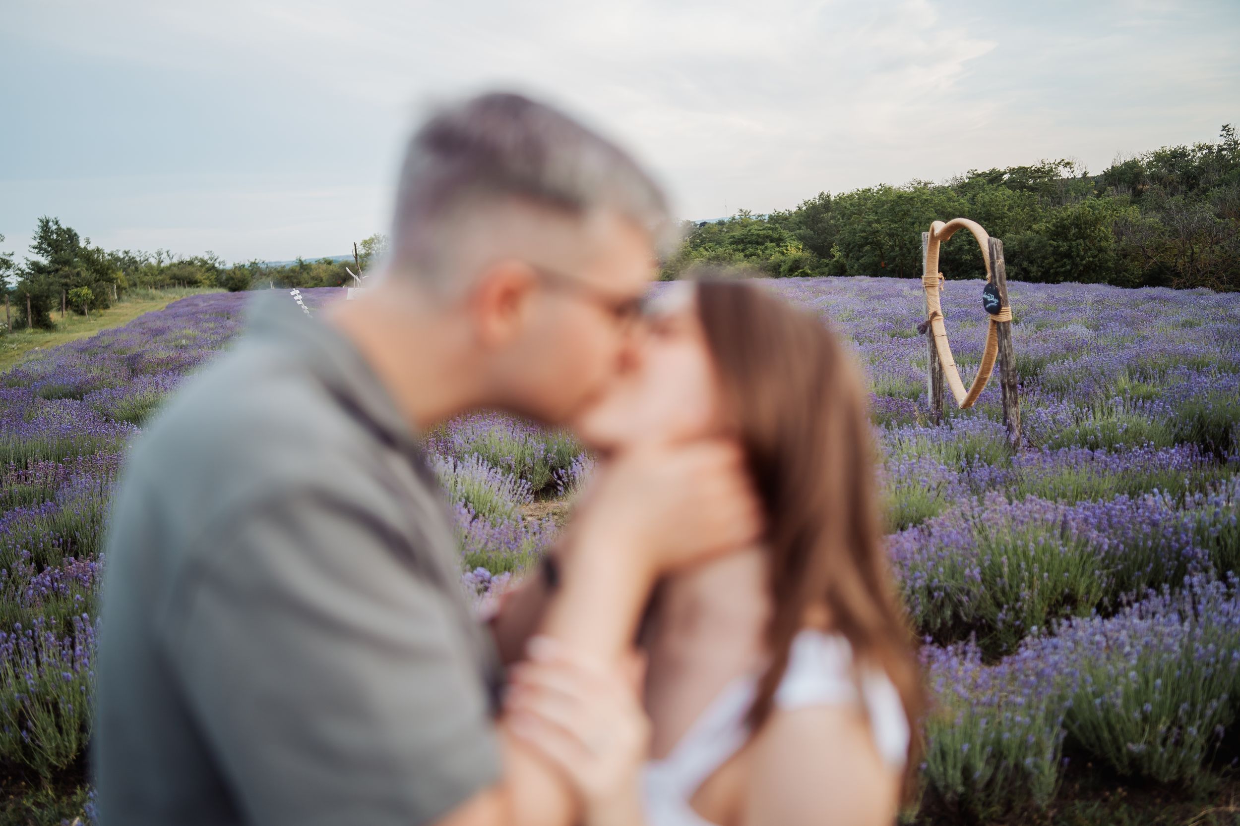 Couple kissing in a blooming lavender field with a blurred figure holding a yoga pose in the background.