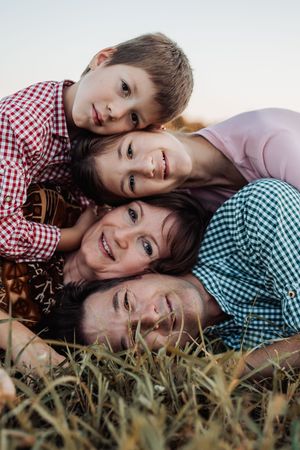 Family of four lying in grass outdoors, smiling and stacking heads, enjoying a sunny day together.
