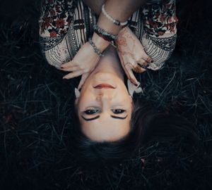Young woman with dark hair and henna tattoos lies on grass, photographed from above in moody, artistic style.