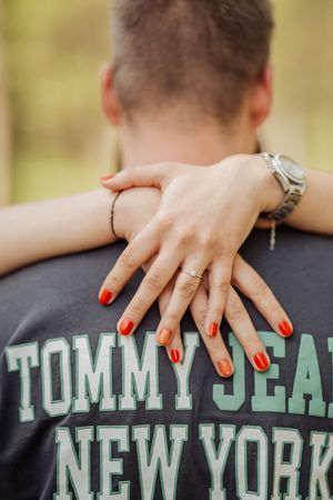 Woman with red nails and silver watch embraces man in Tommy Jean New York shirt, showing engagement ring.
