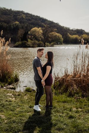 Couple holding hands and gazing at each other by a scenic lake with reeds and tree-covered hills in background.
