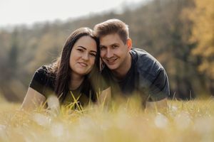 Happy young couple lying in autumn grass field, smiling together outdoors with colorful fall foliage in background.