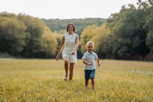 A mother and young boy run joyfully through a sunny green meadow with trees in the background.