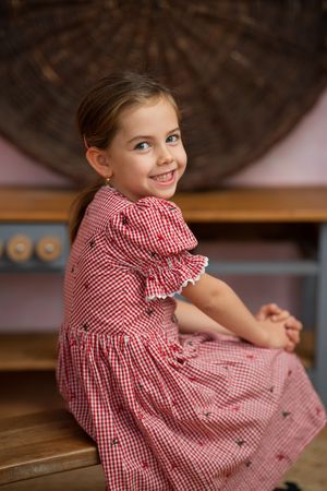 A smiling young girl in a red gingham dress sits on a wooden bench, looking over her shoulder with bright blue eyes.