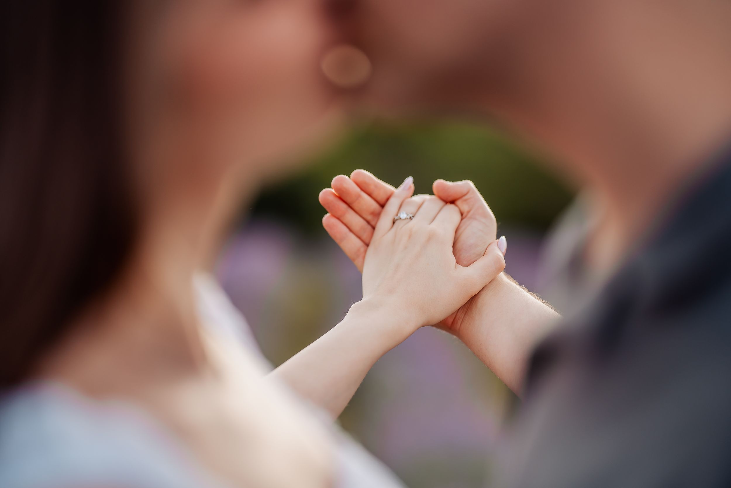 Couple holding hands tenderly while sharing a kiss, with a blurred bokeh background of greenery.