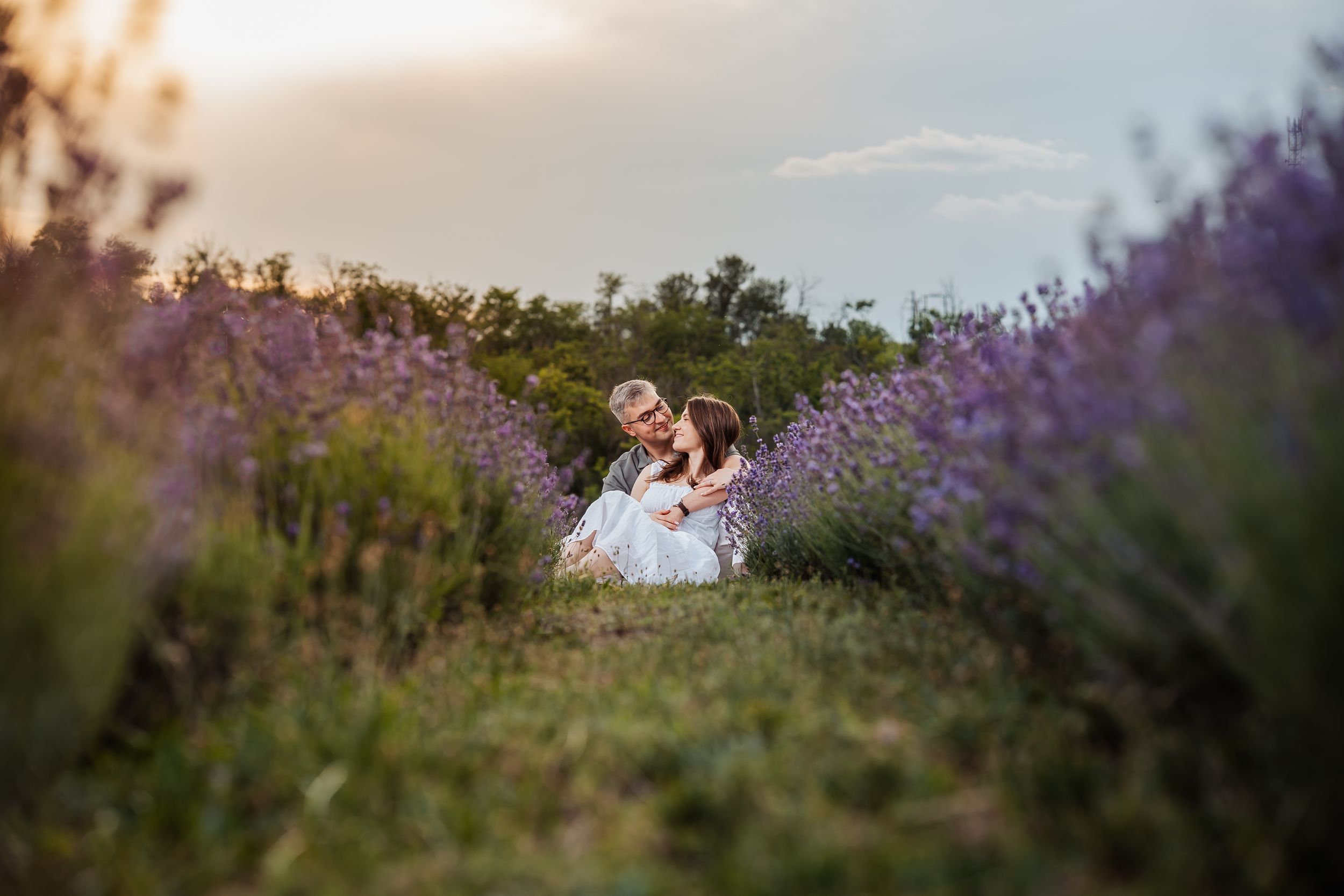 Couple sitting together in a blooming lavender field at sunset, surrounded by purple flowers and lush greenery.