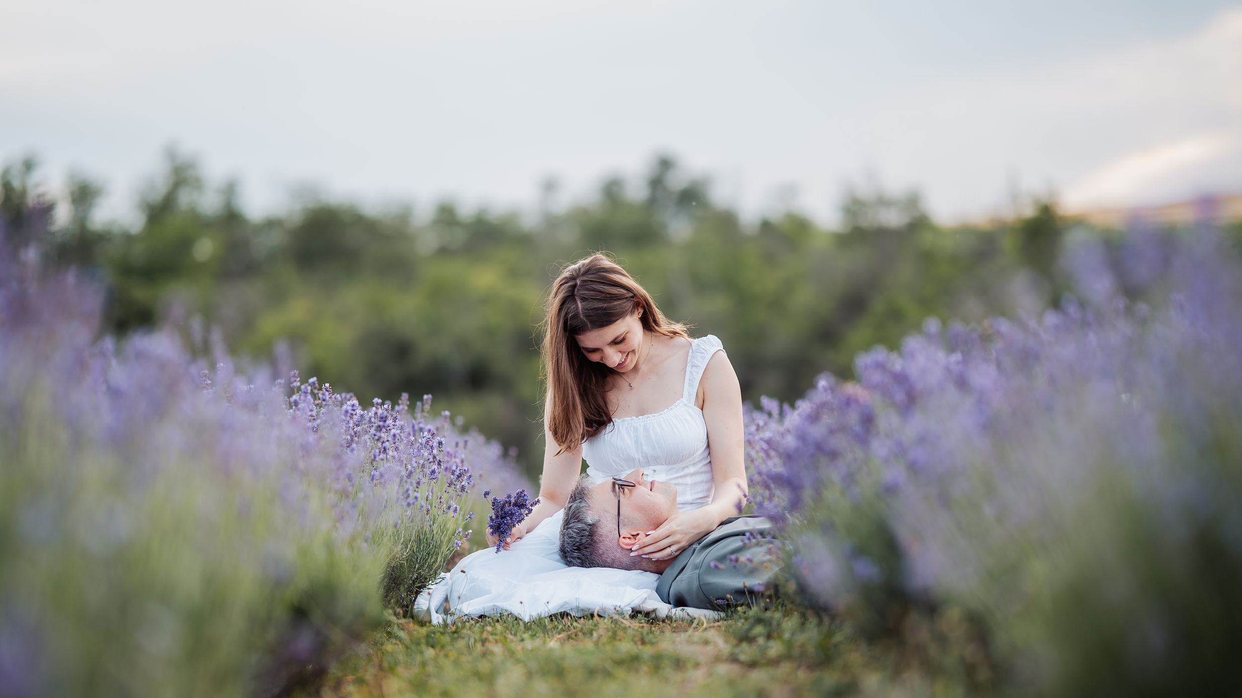 Pregnant woman in white dress sitting in blooming lavender field, cradling her baby bump outdoors.