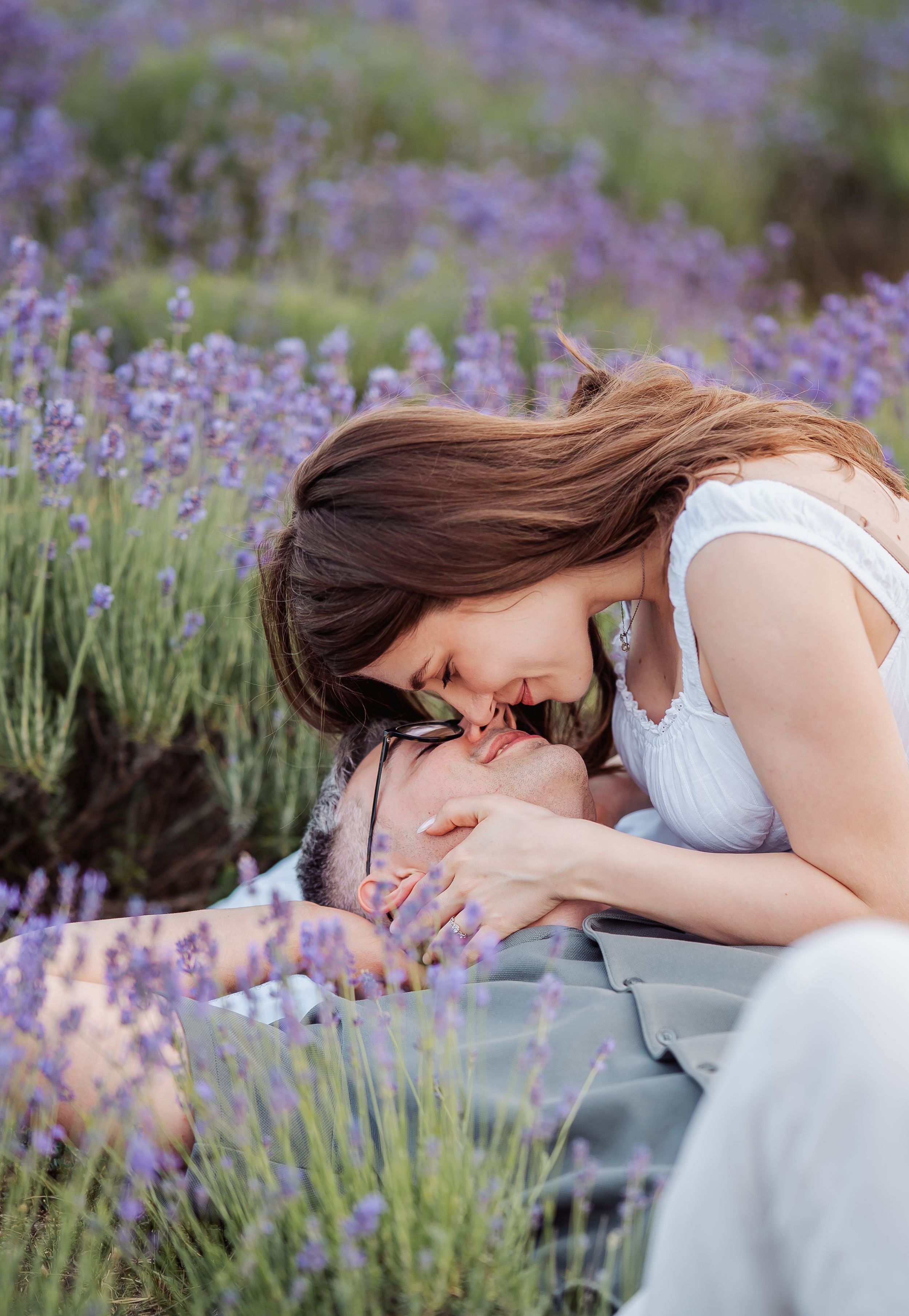 A couple shares a tender romantic moment in a blooming purple lavender field, with the woman leaning down to kiss the man.