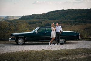 Couple posing beside a classic dark green Cadillac on a scenic overlook with rolling hills in the background.