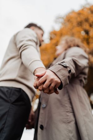 Couple holding hands in an autumn park, wearing beige trench coats, with golden foliage in the background.