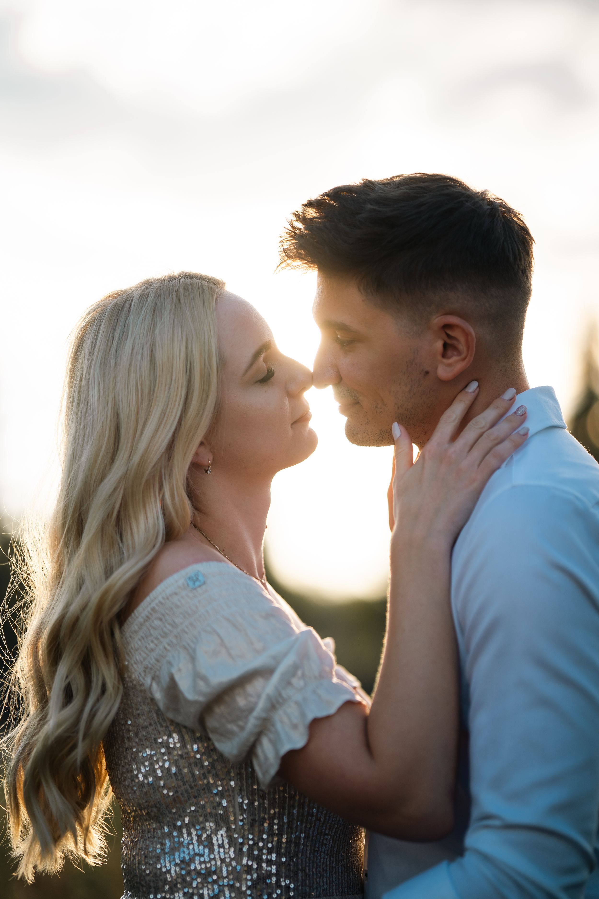 A couple sharing a romantic almost-kiss outdoors, bathed in warm golden sunset light, woman in sequined top with long blonde hair.