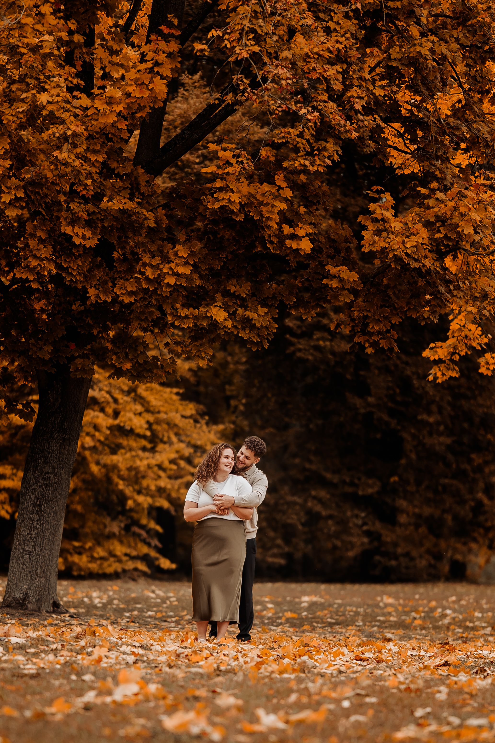 Couple embracing in autumn park with vibrant orange foliage, pregnant woman in olive skirt and white top.