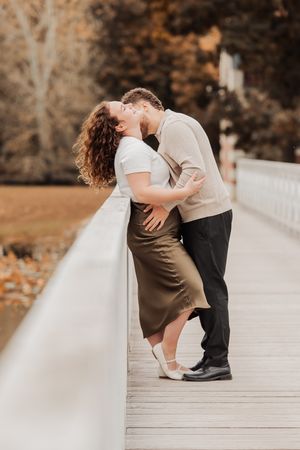 Couple sharing a romantic kiss on a white wooden bridge surrounded by autumn foliage in a park.