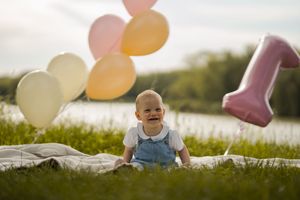 Baby smiling outdoors on grass with colorful balloons and a pink number one balloon celebrating their first birthday.