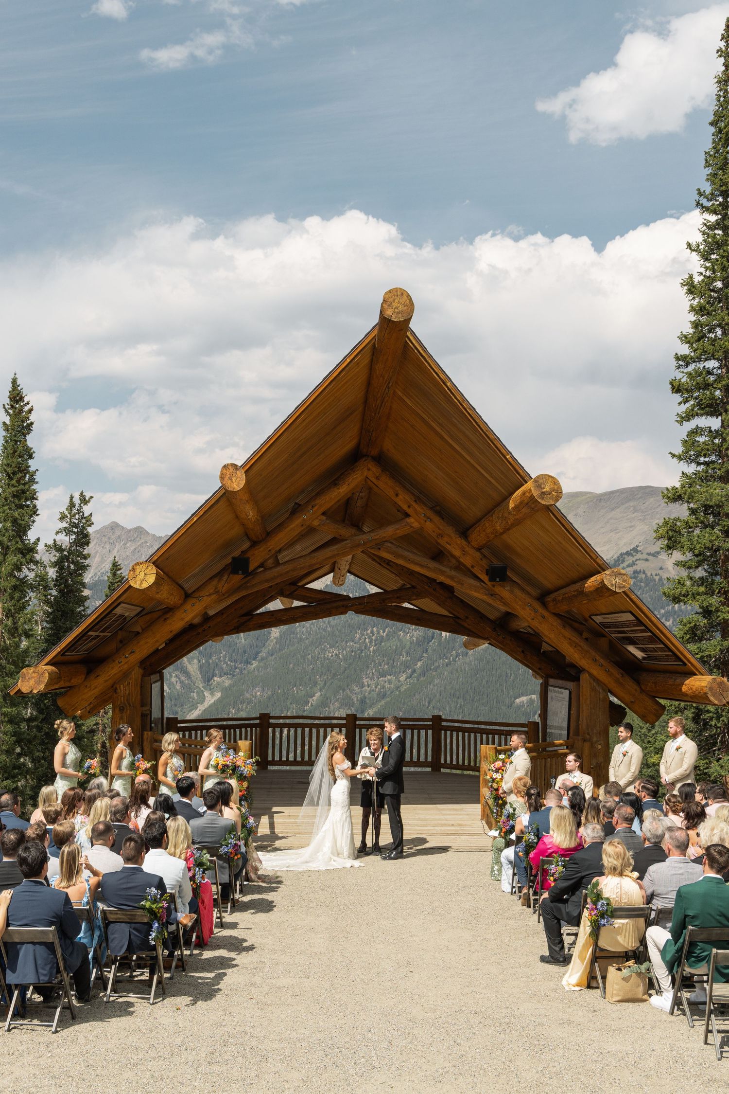 Outdoor mountain wedding ceremony under a rustic log timber frame pavilion with guests seated and scenic peaks backdrop.