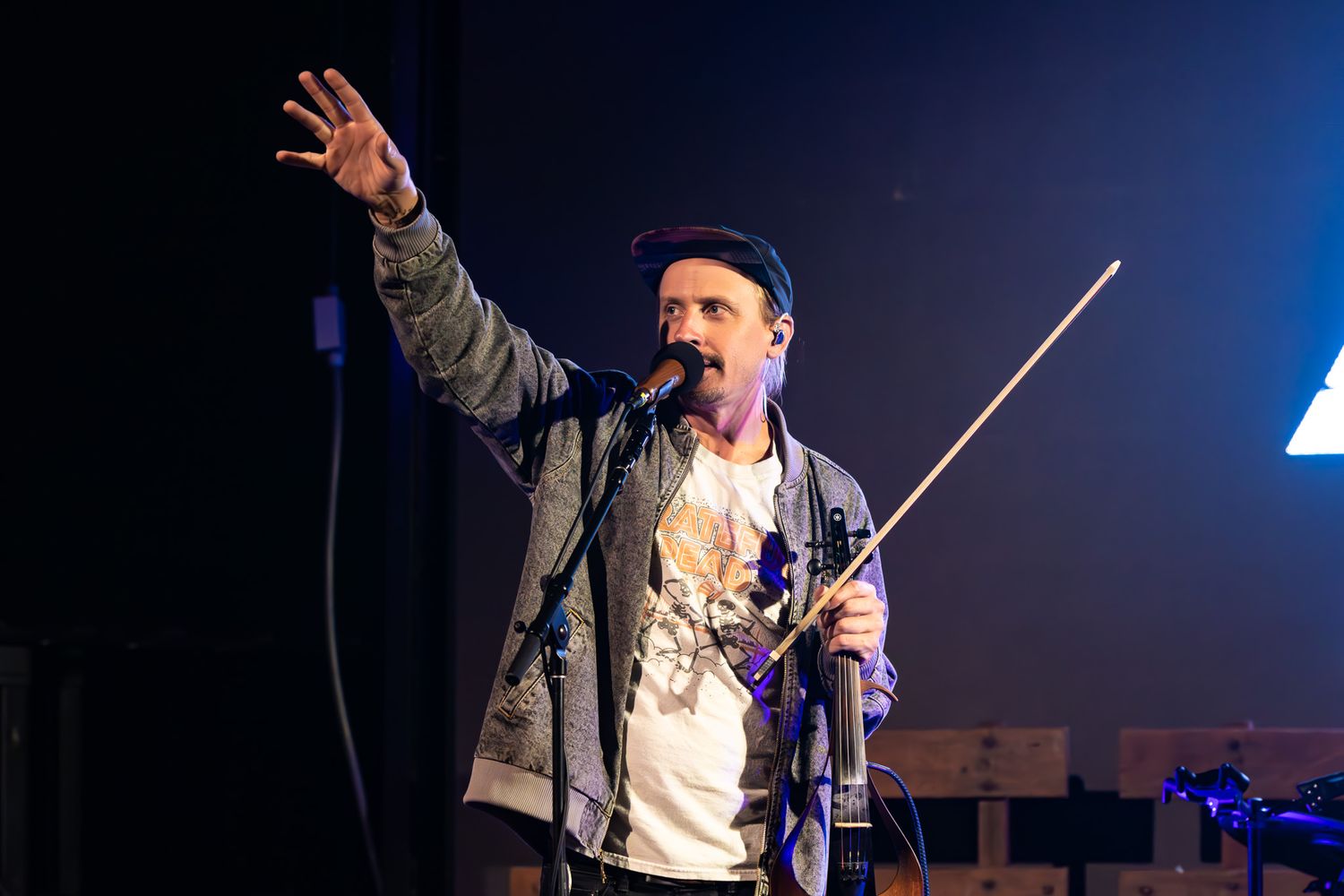 Male performer singing on stage, raising hand, holding conductor baton, wearing denim jacket and blue cap.