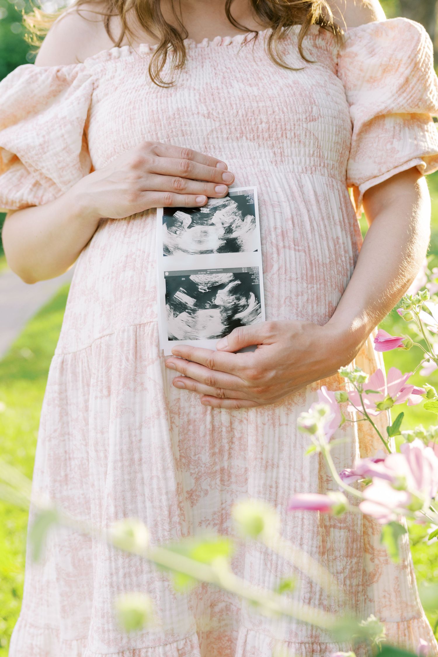 Pregnant woman in pink dress holding ultrasound photos against her baby bump outdoors among flowers.