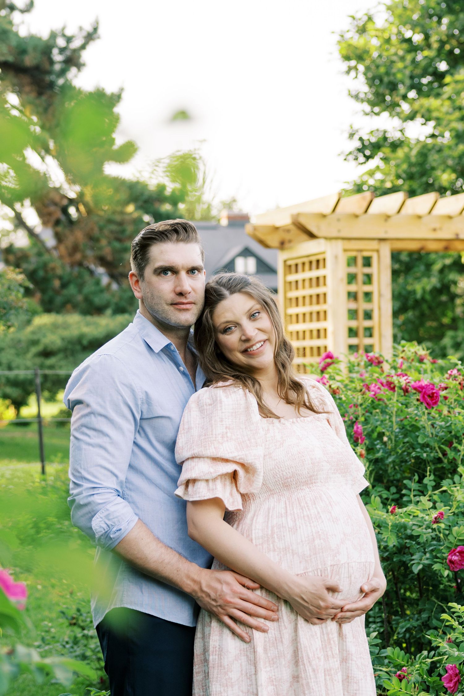 Pregnant couple posing in a lush garden with roses, woman in pink dress cradling her baby bump.