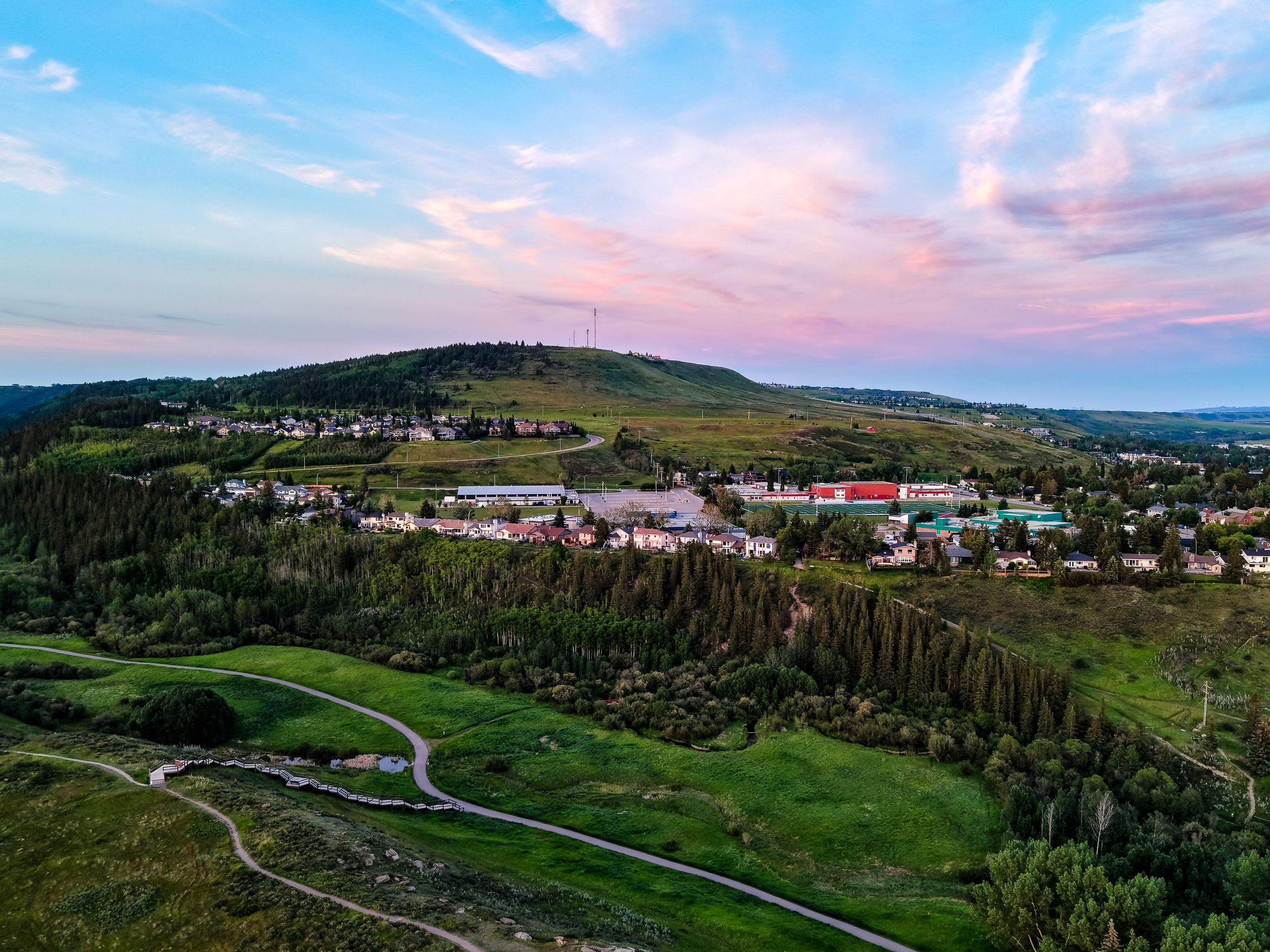 Aerial drone view of a lush green valley town nestled among rolling hills at sunset with colorful pink sky.