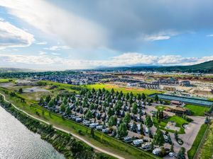 Aerial view of a riverside RV campground surrounded by trees, with a town and mountains in the background.